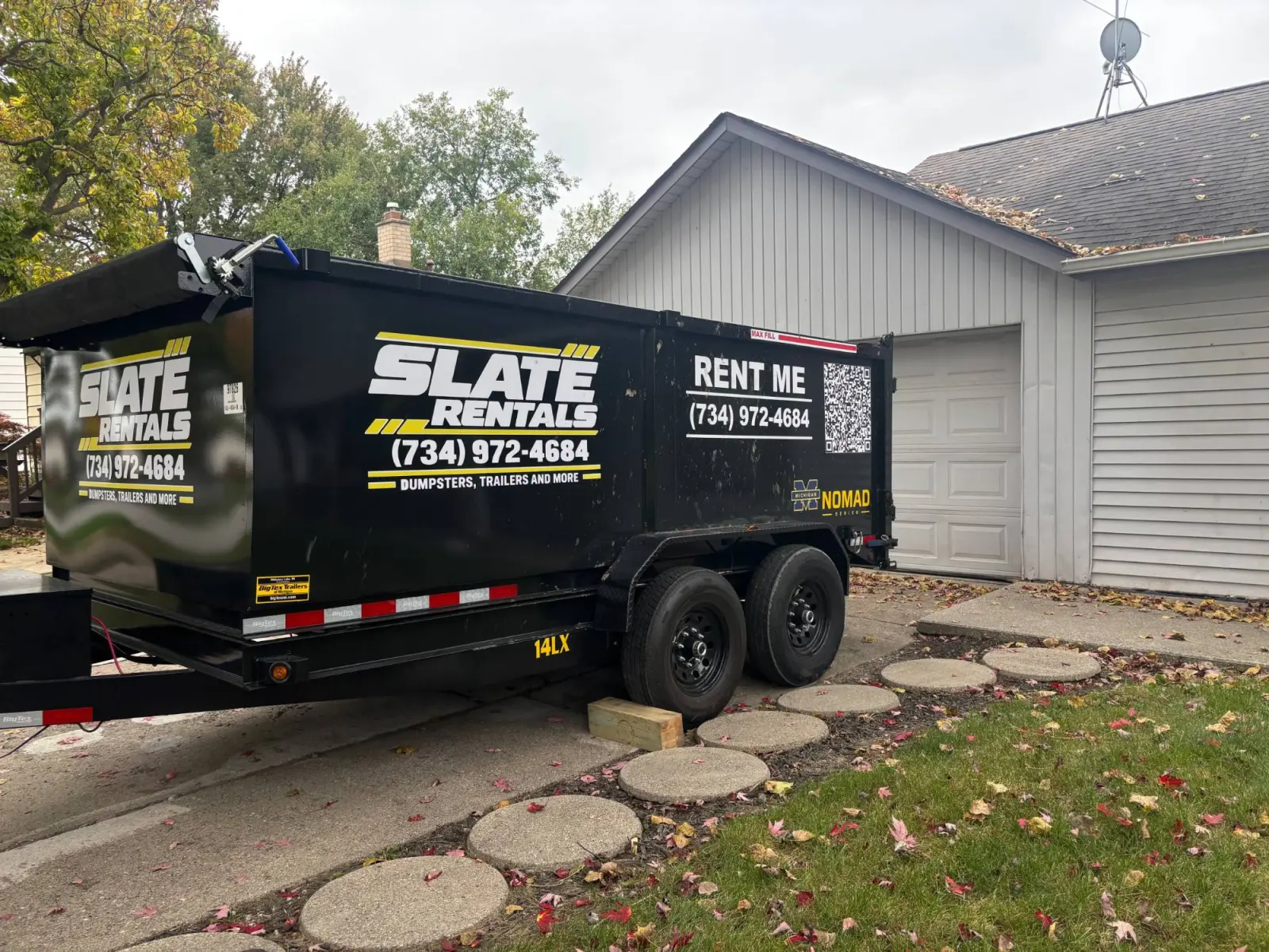 Slate Rentals dump trailer parked in a residential driveway beside a garage, with a wooden board under the tires.